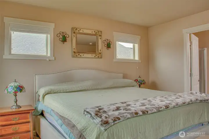 PRIMARY BEDROOM: Two West-Facing Windows with 2" Horizontal Vinyl Blinds; Comfortably Fits King Bed and Furniture; Primary Bathroom Entry Visible on Right, Carpet Flooring.