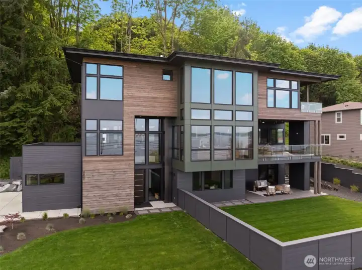 Luxury modern home with expansive water views and upscale contemporary design. Covered patios and view deck with sleek glass railing offer refined outdoor living, complemented by a manicured lawn.