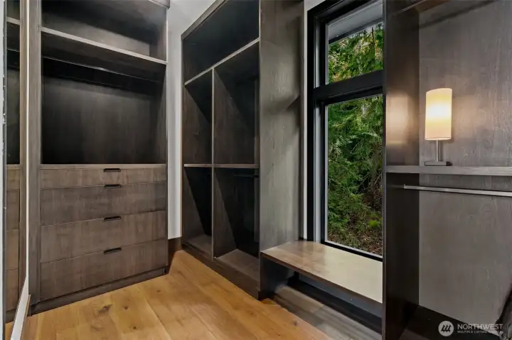 Custom walk-in closet with built-in shelving, drawers, and hanging space, plus a window seat offering a pleasant wooded view.