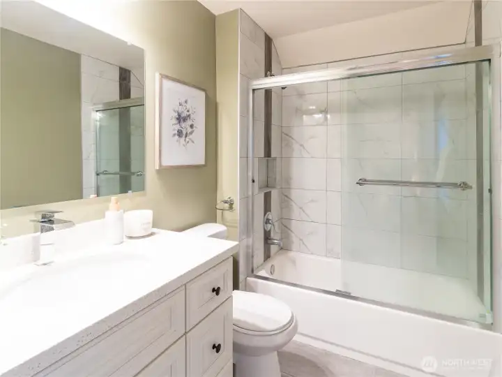 Primary bathroom boasts large vanity, newer toilet, newer tub with tiled tub surround, newer shower doors, and extra nook with storage rack for towels or reimagine the space with your very own personal touch!
