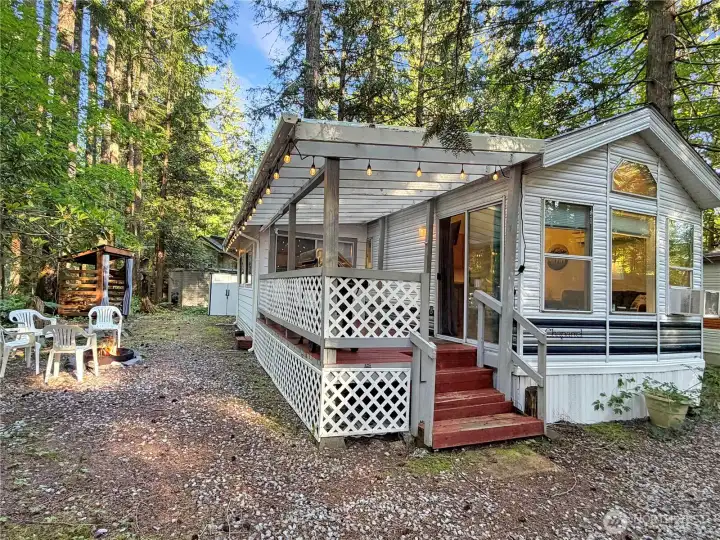 Park model with covered porch, fire pit area, wood shed and two storage sheds in the rear.
