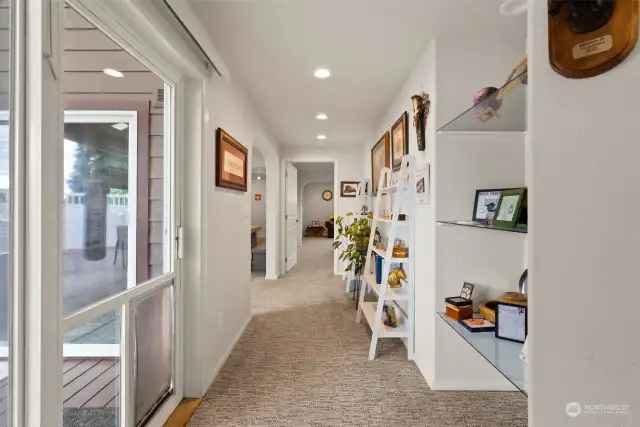 Wide, bright hallway, illuminated by natural light, features built-in glass shelving for displaying art or collectibles, enhancing the open and airy feel. Leading into a spacious primary suite, media/game room, and provides easy access to the hot tub deck, seamlessly connecting key living areas with style and functionality.
