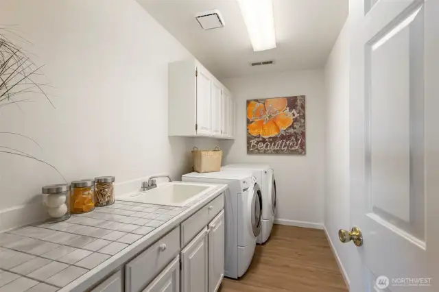 Charming and functional laundry room with a deep utility sink, tile countertop, upper cabinets for storage, and side-by-side washer and dryer.