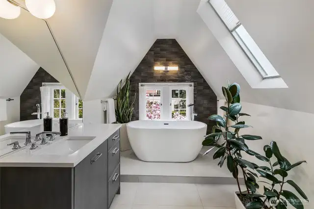 Tucked beneath the gables of the Tower Suite, this En Suite Bath is a spa-like aerie. A freestanding soaking tub rests beneath a skylight and arched stone surround, flanked by greenery and light—an architectural composition of serenity and strength.