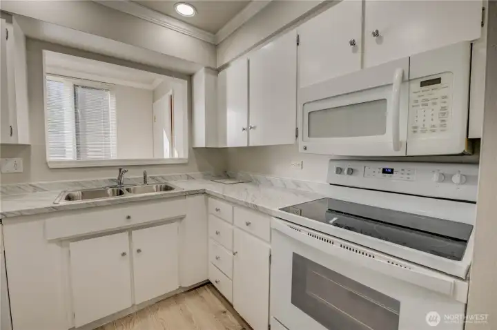 Refreshed kitchen with new LVP flooring and updated countertops with grey veining. Shaker-style cabinets with silver knob finishes.  White electric glass stove stop with over-the-range matching white microwave. Pass-through window looking out to window bringing extra light into the kitchen. Laundry room situated in on the other side of kitchen where pass-thru leads to.