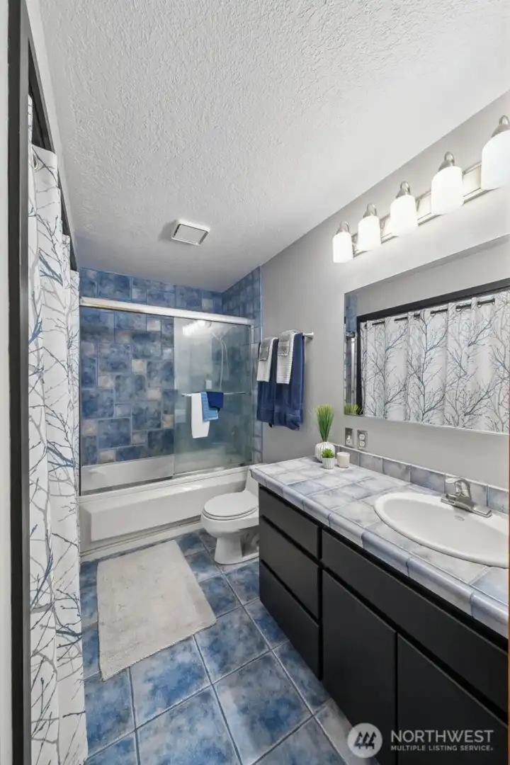 The shared main bathroom offers a large vanity and mirror, functional, durable tile finishes, a glass shower door above a full tub, updated light fixtures and a laundry closet.