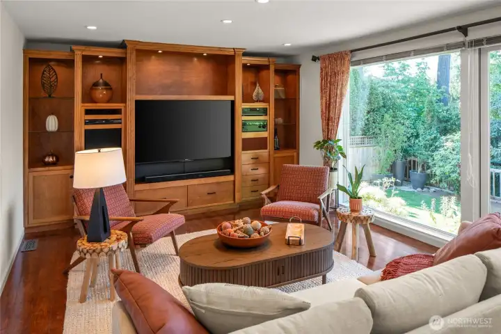 Great room open to kitchen with built-in entertainment center, hardwood floors, and glass wall offering pleasant backyard views