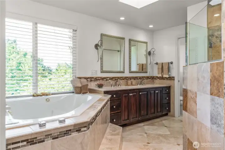 Primary suite bathroom features abundant travertine, a soaking tub with views, double vanities, and a walk-in shower