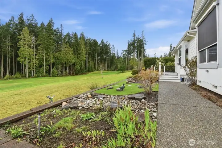 Looking from the East toward the Tee to the west you'll take in the Garden, Water Feature, Astro Turf, and walk path that follows from behind alongside the home to the driveway.
