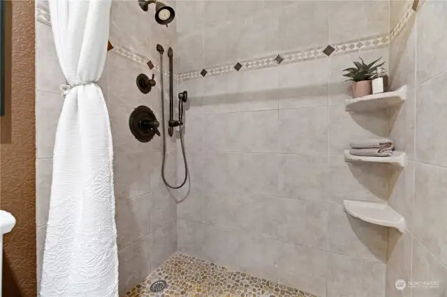 The Primary Bath shower features elegant tile work, a luxurious rainfall showerhead, and convenient built-in shelves for a spa-like experience. Its pebble stone floor adds a touch of natural beauty and provides a relaxing foot massage with every use.