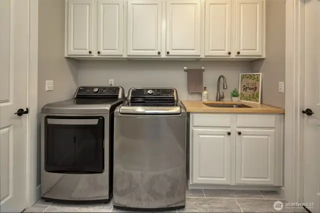 Crisp and functional, this laundry room offers sleek stainless steel appliances, ample upper and lower cabinetry, a butcher block countertop with deep utility sink, and stylish tile flooring—designed for both practicality and charm.