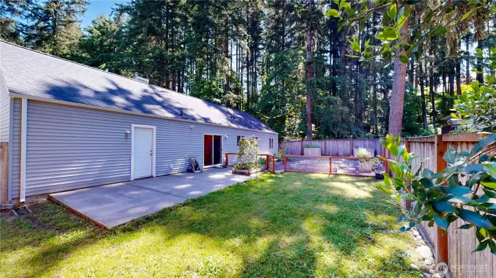 Fully fenced backyard features a spacious concrete patio, level lawn, and a dedicated garden setup with raised beds.