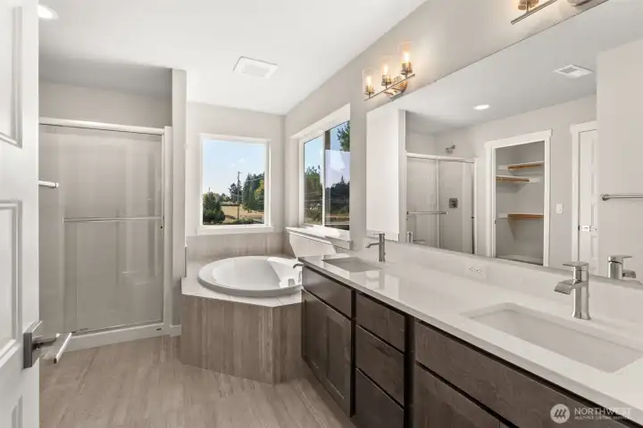 Five-piece primary bath features dual vanity with quartz counters, tile flooring, and tile deck around deep, soaking tub. Walk-in shower features a bench and sliding glass door with brushed nickel finish.