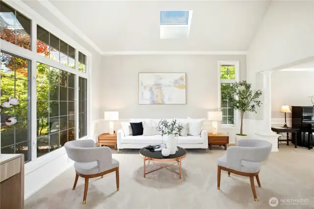 Natural light pours into the elegant living room through grand windows framed with crisp white trim, highlighting the vaulted ceilings and stunning skylight above. Neutral tones and soft carpeted floors set the stage for a serene and welcoming atmosphere, perfectly complemented by wainscoting details and polished crown molding.