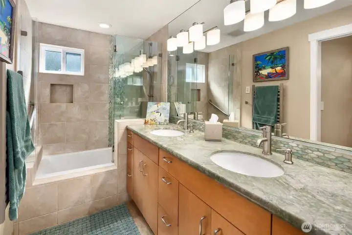 The primary bath: double sinks on a green-veined vanity, a soaking tub and glass shower, and a wrap of stone-look tile.