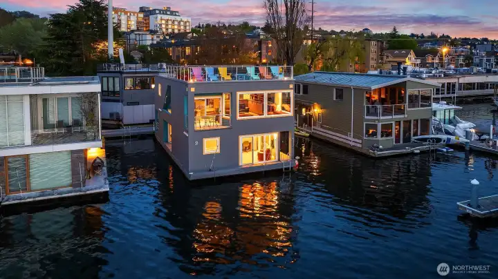 The home glowing from the water at dusk: warm interiors visible through every pane, rooftop deck lit above, and a purple sky folding over the city.