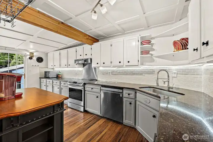 Wrap around galley kitchen with corner sink and open shelving above sink. Wall to wall upper cabinets, and two-toned cabinetry. White subway tile blacksplash wraps entire kitchen wall. Stainless appliances, all staying with house. Tracking lighting, exposed beam, and ceiling mounted pots/pans storage rack opening up extra storage space in cabinets.