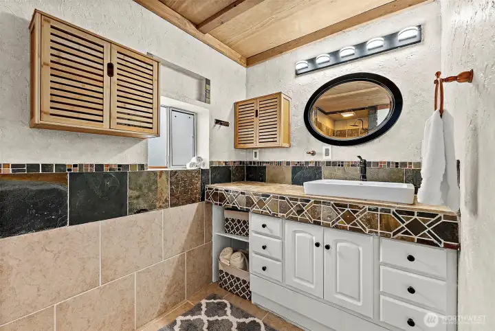 Vanity nook of bathroom opposite the shower features ceramic tile flooring and backsplash. Vanity features a custom mosaic tile top, open shelving and multiple drawers for optimal storage space. Wood paneled ceiling.