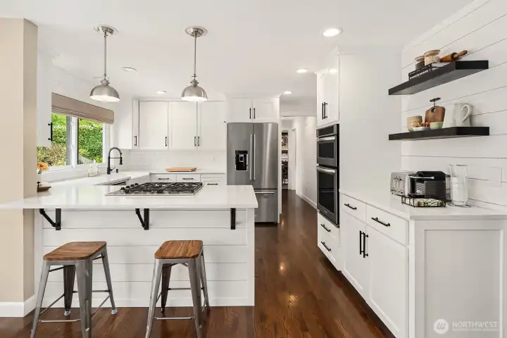 This angle showcases the kitchen's generous layout and ample storage, with open shelving, bar seating at the island, and a wall of windows keeping the space bright and connected to the outdoors.