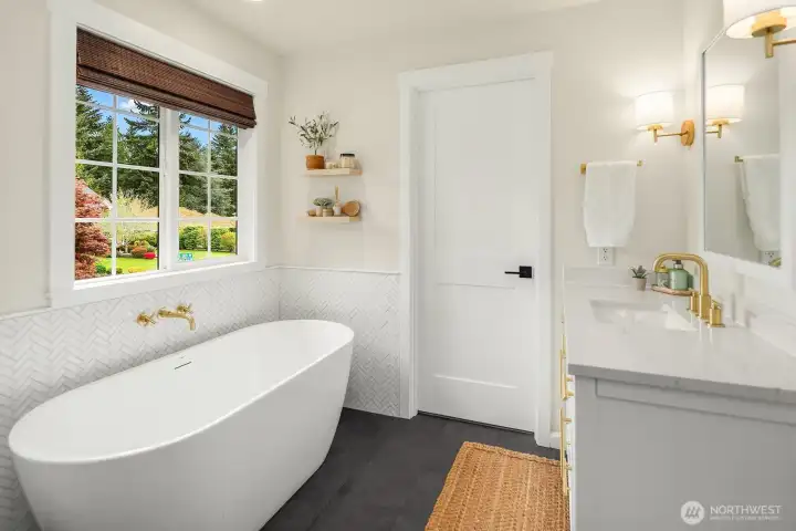 A bright window floods the primary bath with natural light, highlighting the elegant freestanding soaking tub and refined brass fixtures that make this space feel like a true private spa.