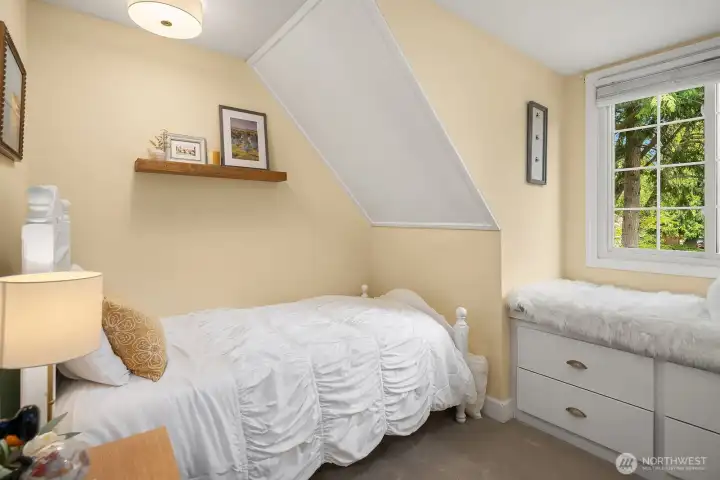 This cheerful upper bedroom features charming roofline architecture, a bright corner window, and built-in storage — a sweet and functional space that works beautifully as a child's room or cozy guest retreat.