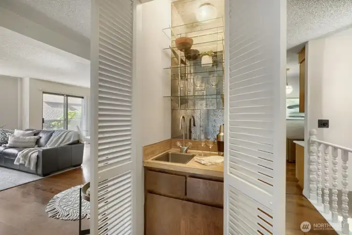 Charming Wet Bar Off Living Room    Perfectly tucked behind louvered doors, this stylish wet bar adds a touch of sophistication and convenience to the living space. Featuring a mirrored backsplash, glass shelving, and a sleek stainless-steel sink, it’s ideal for entertaining or enjoying a quiet evening drink. The warm wood cabinetry complements the engineered hardwood floors and open layout, while its location just off the living room makes it both functional and inviting.