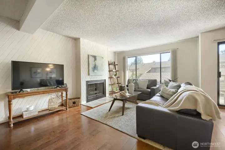 Bright Living Room with Fireplace and Balcony Access    This spacious living room offers a warm, inviting atmosphere with hardwood floors, neutral tones, and abundant natural light from large windows and sliding glass doors. A wood-paneled accent wall frames the classic fireplace, creating a cozy focal point ideal for relaxing or entertaining. The open layout flows seamlessly to the balcony, providing a perfect indoor-outdoor connection and views of the surrounding community.