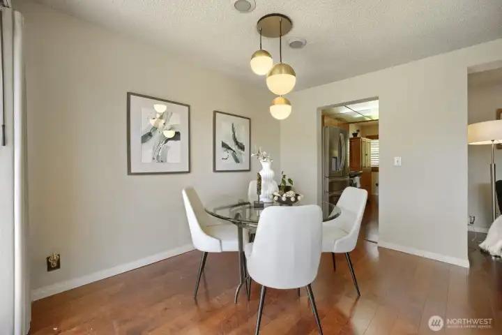 Bright Dining Area Off Kitchen and Deck    This stylish dining space sits just off the kitchen and opens toward the deck, creating an effortless indoor-outdoor flow perfect for entertaining. Warm engineered hardwood floors complement the soft neutral palette, while a modern glass-top table and sculptural pendant lighting add a touch of sophistication. Framed abstract art and natural light enhance the inviting atmosphere, making this the ideal spot for everyday meals or relaxed gatherings.