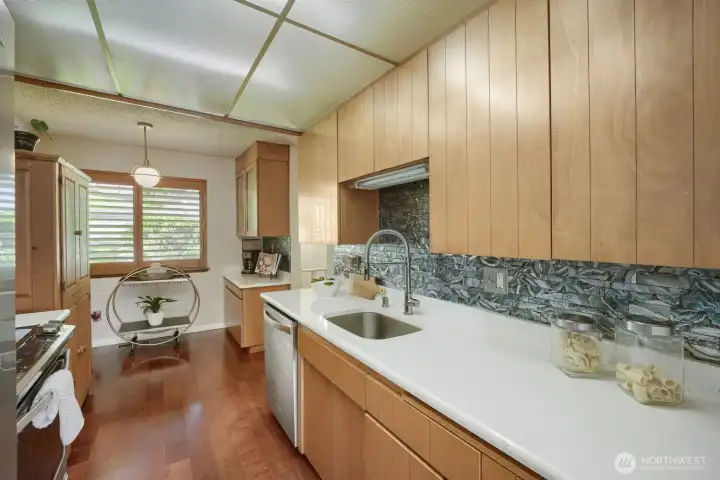 Inviting Kitchen with Stand-Alone Pantry and Coffee Bar    This thoughtfully designed kitchen blends warmth and functionality with light wood cabinetry, white solid-surface countertops, and a stunning textured tile backsplash that adds depth and character. A stand-alone pantry provides extra storage and style, while the dedicated coffee bar creates a perfect morning ritual space. Stainless-steel appliances and a sleek undermount sink complement the modern aesthetic, and engineered hardwood floors flow seamlessly throughout. Natural light from the large window enhances the bright, welcoming atmosphere—ideal for both everyday cooking and entertaining.