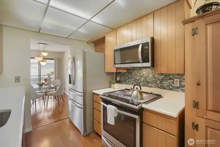 Bright Kitchen with Modern Touches    This inviting kitchen blends warmth and functionality with natural wood cabinetry, stainless-steel appliances, and a stylish patterned backsplash that adds character. The smooth white countertops and updated lighting create a clean, contemporary feel. A pass-through layout opens to the dining area, where large sliding doors fill the space with natural light—perfect for entertaining or everyday living.