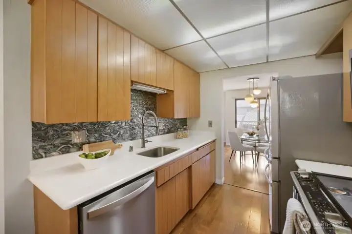 Contemporary Kitchen with Warm Wood Accents    This thoughtfully designed kitchen combines style and functionality with light wood cabinetry, white solid-surface countertops, and a striking textured tile backsplash that adds depth and character. Stainless-steel appliances, including a refrigerator, dishwasher, and range, bring a modern touch, while the engineered hardwood floors continue seamlessly from the main living area. The open layout connects directly to the dining space, creating an inviting flow for cooking and entertaining. Clean lines, natural light, and timeless finishes make this kitchen both elegant and efficient.