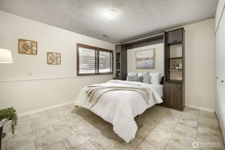 Lower-Level Bedroom with Heated Floors    This beautifully finished lower-level bedroom offers comfort and style with heated tile floors that provide warmth year-round. The space features a neutral palette, textured ceiling, and a large window with wood shutters that fill the room with natural light. Built-in shelving and cabinetry frame the bed, adding both function and elegance. Perfect as a guest suite, home office, or private retreat, this room combines modern convenience with cozy sophistication.