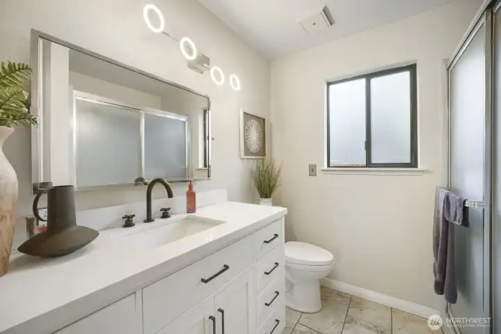 Modern Bathroom with Sleek Finishes    This beautifully updated bathroom features a white vanity with quartz countertop, accented by matte black fixtures and contemporary hardware. A large framed mirror and ring-style LED lighting create a bright, stylish atmosphere. The walk-in shower with frosted glass doors adds privacy and function, while the neutral tile flooring and soft wall tones provide a clean, timeless look. Thoughtful details and natural light make this space both elegant and inviting.