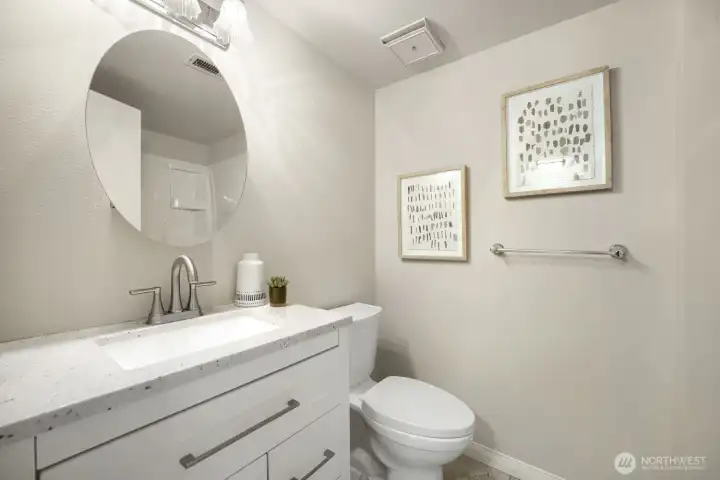 Updated Bathroom with Modern Touches    This stylish bathroom features a white vanity with quartz countertop, brushed-nickel fixtures, and a round mirror accented by a contemporary light fixture. Neutral tones and tile flooring create a clean, calming atmosphere, while framed artwork adds a touch of sophistication. The space is bright, functional, and thoughtfully designed—perfect for everyday comfort or guest use.