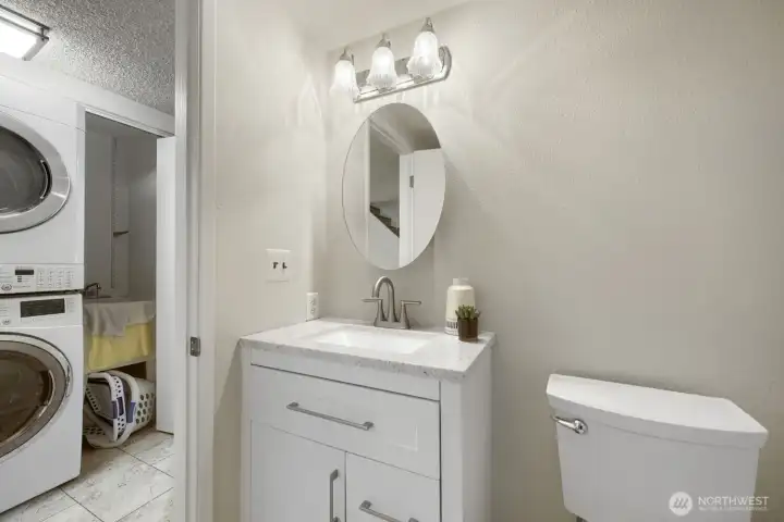 Downstairs Bathroom Adjacent to Bedroom    This refreshed downstairs bathroom offers modern convenience and clean design. It features a white vanity with quartz countertop, brushed-nickel fixtures, and a round mirror accented by a stylish three-light fixture. Neutral tones and tile flooring create a bright, cohesive look that complements the adjacent bedroom. Located next to the laundry area, this space combines practicality with contemporary charm—perfect for guests or everyday use.