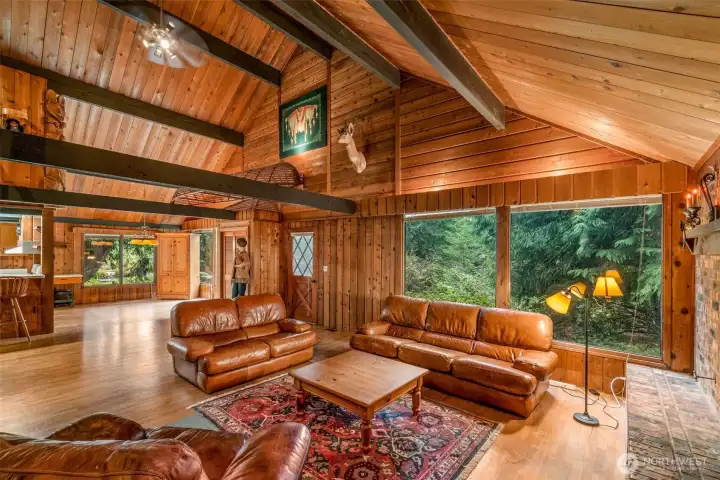 From the stairs, the lodge stretches wide and tall—cedar ceiling soaring above, beams drawing the eye across the space. Huge windows frame the trees in a living mural, while the dining room nestles into the back corner. Overhead, a deer trophy keeps quiet watch, joined by striking Native American art and a totem that add depth and character to the scene.