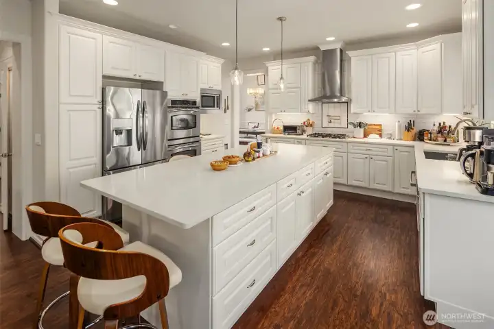 A larger and updated kitchen with pull out shelving, soft close hinges, pantry space, pendant lighting over the huge island, breakfast bar space, Quartz counters, French door refrigerator, dual wall ovens, microwave oven, and a large deep stainless stell sink