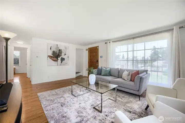 Bright and welcoming, this living room blends modern comfort with timeless charm. Sunlight pours through the large picture window, illuminating the warm hardwood floors and crisp white walls. A contemporary gray sectional anchors the space, accented by soft-hued pillows that add a touch of color and sophistication. The glass coffee table and abstract artwork create a polished, gallery-like feel, while the wood front door adds a hint of mid-century character. Perfect for relaxing or entertaining, this airy space offers a seamless balance of style and warmth. Bright and welcoming, this living room blends modern comfort with timeless charm. Sunlight pours through the large picture window, illuminating the warm hardwood floors and crisp white walls. A contemporary gray sectional anchors the space, accented by soft-hued pillows that add a touch of color and sophistication. The glass coffee table and abstract artwork create a polished, gallery-like feel, while the wood front door adds a hint of mid-century character. Perfect for relaxing or entertaining, this airy space offers a seamless balance of style and warmth.