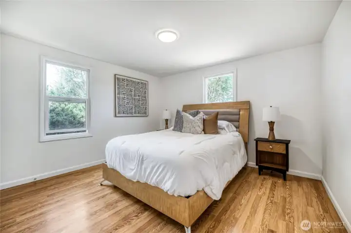 The primary bedroom offers a bright and restful retreat with clean lines and warm wood flooring. Two large windows fill the space with natural light and frame views of lush greenery outside. The neutral palette and simple design create a calm, modern atmosphere, while the generous layout easily accommodates a full bedroom set. Perfect for relaxation, this space blends comfort and style beautifully. The primary bedroom offers a bright and restful retreat with clean lines and warm wood flooring. Two large windows fill the space with natural light and frame views of lush greenery outside. The neutral palette and simple design create a calm, modern atmosphere, while the generous layout easily accommodates a full bedroom set. Perfect for relaxation, this space blends comfort and style beautifully.
