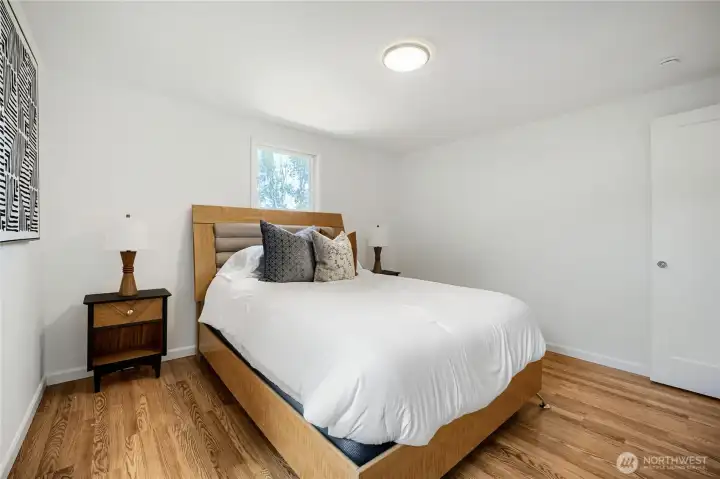 The primary bedroom offers a bright and restful retreat with clean lines and warm wood flooring. Two large windows fill the space with natural light and frame views of lush greenery outside. The neutral palette and simple design create a calm, modern atmosphere, while the generous layout easily accommodates a full bedroom set. Perfect for relaxation, this space blends comfort and style beautifully. The primary bedroom offers a bright and restful retreat with clean lines and warm wood flooring. Two large windows fill the space with natural light and frame views of lush greenery outside. The neutral palette and simple design create a calm, modern atmosphere, while the generous layout easily accommodates a full bedroom set. Perfect for relaxation, this space blends comfort and style beautifully.