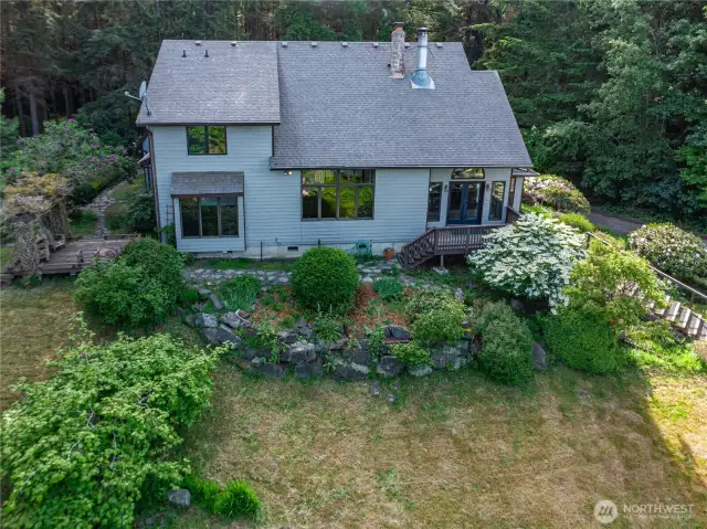 The east side of the home features stone pathways, a deck, and steps leading to a grassy parking area and gardens. The sliding glass door on the right opens to the kitchen, while the primary bedroom is located behind the lower left window.