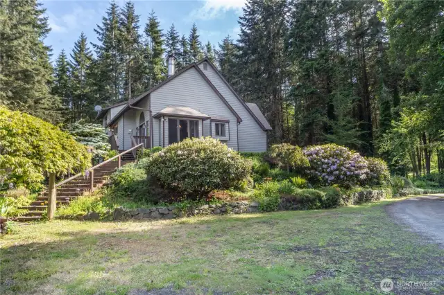 The north side of the house has a grassy area for parking, with a stairway up to the deck and pathway to the front door. Private and peaceful, and a gardener's paradise. The window in the photo is the breakfast nook in the kitchen.