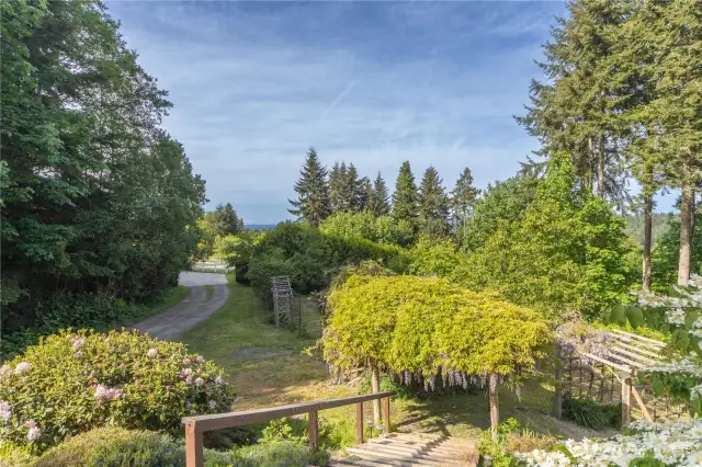 The peek-a-boo water and mountain views from the north side of the home. Looking down to the grassy parking area, and a fenced vegetable garden and storage shed behind the wisterial.