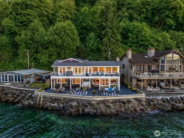 From the water at dusk, the waterfront home glows like a quiet jewel nestled along the shoreline. Wide decks and spacious waterside patio, extend outward. lit with subtle recessed lighting. A picture of peace and quiet luxury- a home that feels both grounded in nature and living in perfect grandeur.