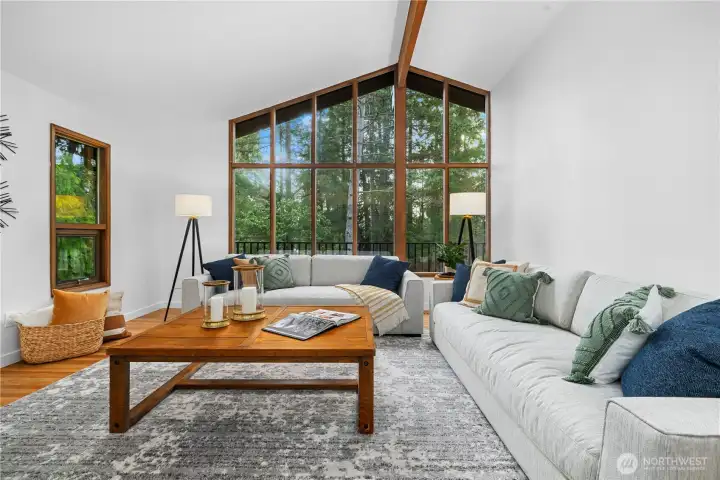 Light-filled living room with vaulted ceilings, exposed beam detail, and dramatic floor-to-ceiling windows overlooking a private, wooded setting.