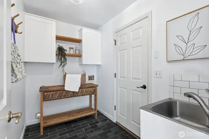 Functional main-floor laundry room with tile flooring, built-in cabinetry, shelving, and a utility sink, conveniently located off the kitchen with direct garage access.