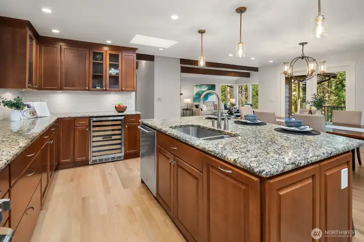 This expansive view of the kitchen showcases the generous layout with abundant counter space, stunning granite surfaces, and seamless flow toward the bright windows overlooking the lush backyard. The warm wood cabinetry, refinished hardwood floors, and updated pendant lighting combine to create a kitchen that is as beautiful as it is practical.