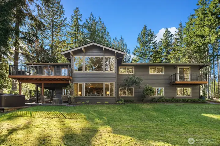 This impressive rear exterior view showcases the home's grand two-story presence, featuring the expansive main level deck above and the lower level patio below, both perfectly positioned to enjoy the beautifully landscaped, sun-drenched backyard. The lush lawn, mature evergreen backdrop, and thoughtful outdoor living spaces make this backyard a true Pacific Northwest paradise.