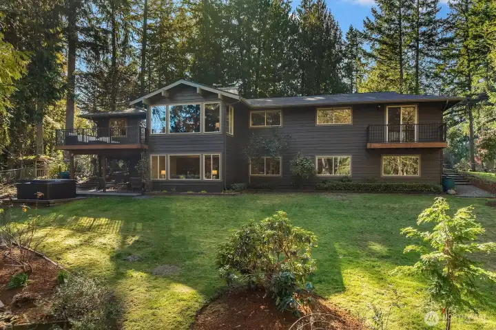This stunning alternate view of the home's rear exterior highlights the beautifully maintained grounds, vibrant mature landscaping, and the home's impressive two-story presence framed perfectly by towering Pacific Northwest evergreens. The lush, manicured lawn, full sprinkler system, and deer-proof fencing come together to create an idyllic and private outdoor sanctuary.