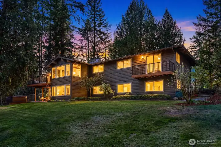 Photo 40 – Twilight Rear Exterior  This stunning twilight view of the home's rear exterior is nothing short of spectacular, with warm light glowing from every window and illuminating the multi-level decks against a gorgeous dusky sky and towering evergreen backdrop. This captivating image perfectly encapsulates the grandeur, warmth, and exceptional lifestyle this remarkable Sammamish home has to offer.